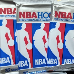 May include: Five unopened packs of NBA Hoops basketball trading cards. The packs are white with blue and red accents. Each pack features a silhouette of a basketball player dribbling a ball. The text "NBA Hoops" is printed on the front of each pack.