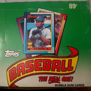 May include: A green Topps baseball bubble gum cards box featuring a Ken Griffey Jr. Mariners card. The box displays the text BASEBALL THE REAL ONE! and 89c. The card shows a player in a light blue uniform holding a bat with the name KEN GRIFFEY, JR.