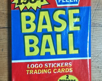 May include: A red, white, and blue 1984 Fleer Baseball trading card pack with a sticker and 15 cards. The pack features a yellow starburst with the year 1984, a blue and yellow baseball logo, and a red and white Fleer logo. The pack also includes a sticker and 15 cards with two photos on each card.