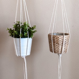Puede incluir: Dos maceteros colgantes de macramé con anillos de madera, uno con una maceta blanca con una planta verde y el otro con una cesta de mimbre tejida.