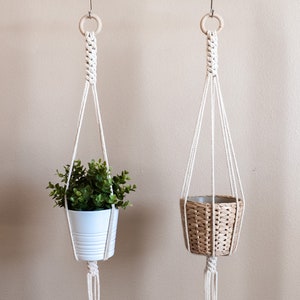 Puede incluir: Dos colgadores de plantas de macramé suspendidos de una barra blanca. Uno sostiene una maceta blanca con follaje verde, el otro una cesta tejida. Los colgadores son de cuerda blanquecina, con aros de madera en la parte superior.