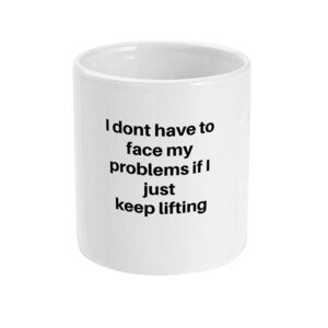 Könnte beinhalten: Weißer Keramikbecher mit dem Text "I dont have to face my problems if I just keep lifting" in Schwarz. Der Becher ist zylindrisch und hat eine glatte, glänzende Oberfläche. Der Text ist zentriert.