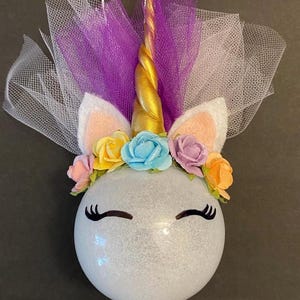 May include: A white glitter ornament with a unicorn face, gold horn, purple tulle, and a flower crown with yellow, blue, pink, and orange flowers.
