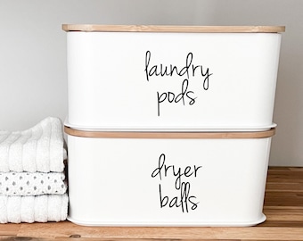 May include: Two white rectangular storage containers with bamboo lids. The top container is labelled "laundry pods" and the bottom container is labelled "dryer balls".