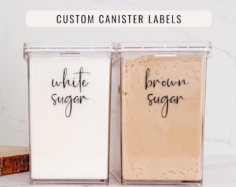 May include: Two clear rectangular plastic canisters with airtight lids. The left canister contains white sugar and the right contains brown sugar. Each canister features black cursive text labels reading white sugar and brown sugar respectively.