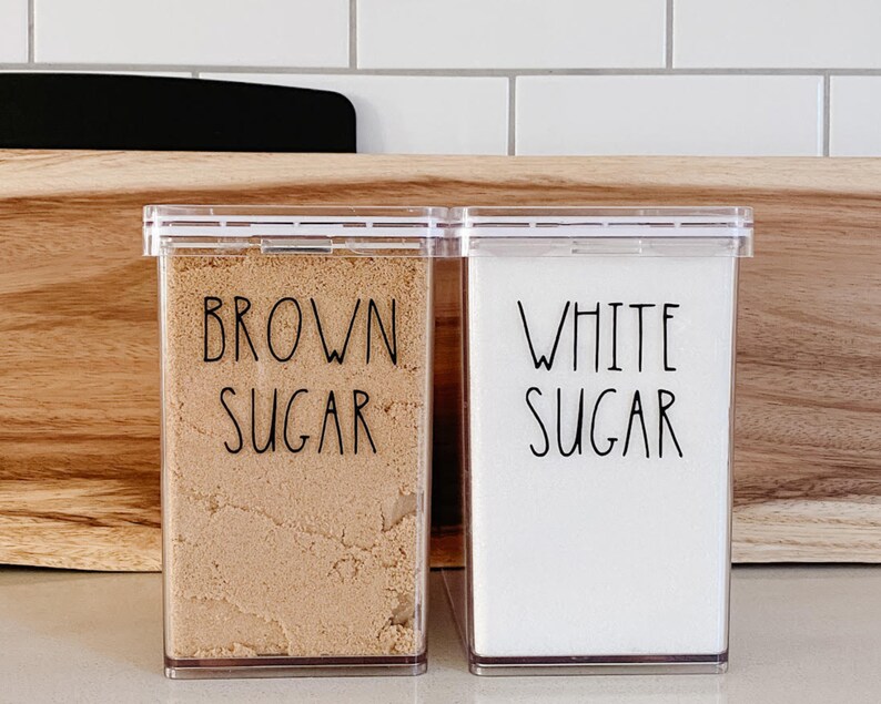 May include: Two clear plastic containers with black labels. The container on the left is filled with brown sugar and labeled "BROWN SUGAR". The container on the right is filled with white sugar and labeled "WHITE SUGAR".