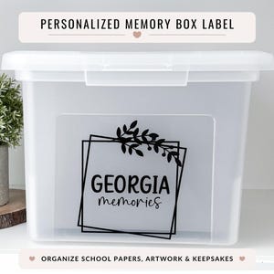 Peut inclure: Une boîte de rangement transparente avec un couvercle blanc. L'étiquette indique "GEORGIA memories" en noir, dans une police stylisée, encadrée d'un motif géométrique noir et d'éléments floraux. L'étiquette supérieure indique "PERSONALIZED MEMORY BOX LABEL".