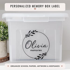 May include: A white plastic memory box with a lid, featuring a personalized label with the name "Olivia" and the word "memories" inside a circular wreath design. The text "PERSONALISED MEMORY BOX LABEL" is above the box.