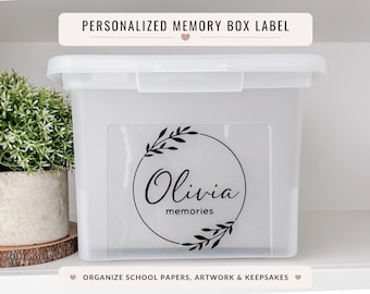 May include: A white plastic memory box with a lid, featuring a personalized label with the name "Olivia" and the word "memories" inside a circular wreath design. The text "PERSONALIZED MEMORY BOX LABEL" is above the box.