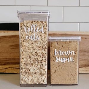 May include: Two clear plastic containers with white labels. The container on the left is filled with rolled oats and labeled "rolled oats". The container on the right is filled with brown sugar and labeled "brown sugar".