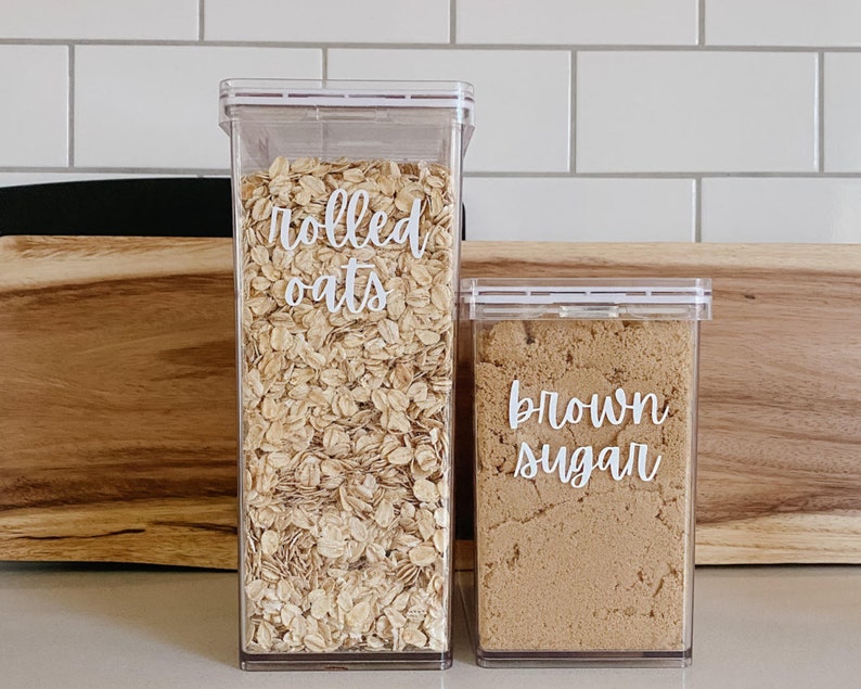 May include: Two clear plastic containers with white labels. The container on the left is filled with rolled oats and labeled "rolled oats". The container on the right is filled with brown sugar and labeled "brown sugar".