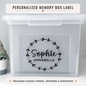 Peut inclure: Une boîte de rangement transparente avec un couvercle, portant l'inscription "PERSONALIZED MEMORY BOX LABEL". La boîte présente une étiquette avec le nom "Sophie Annabelle" dans une couronne florale noire. Le texte "ORGANIZE SCHOOL PAPERS, ARTWORK & KEEPSAKES" est également visible.