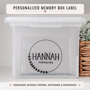 May include: A clear plastic memory box with a label that reads "HANNAH memories" in a black circular design. The box has a white lid and is next to a woven basket with greenery. Text above reads "PERSONALIZED MEMORY BOX LABEL".