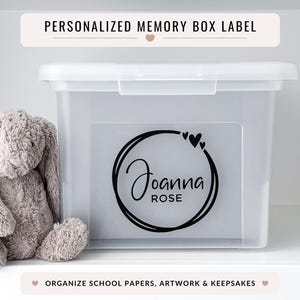 May include: A clear plastic storage box with a white lid and a personalised label. The label features the name "Joanna Rose" in a black script within a circular design with hearts. A plush grey bunny sits beside the box. Text above reads "PERSONALISED MEMORY BOX LABEL".