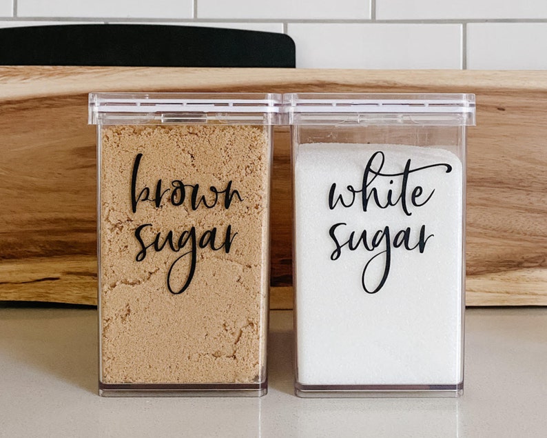 May include: Two clear plastic containers with black labels that say "brown sugar" and "white sugar". The containers are filled with brown sugar and white sugar.