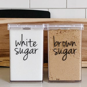 May include: Two clear plastic containers with labels that say "white sugar" and "brown sugar".