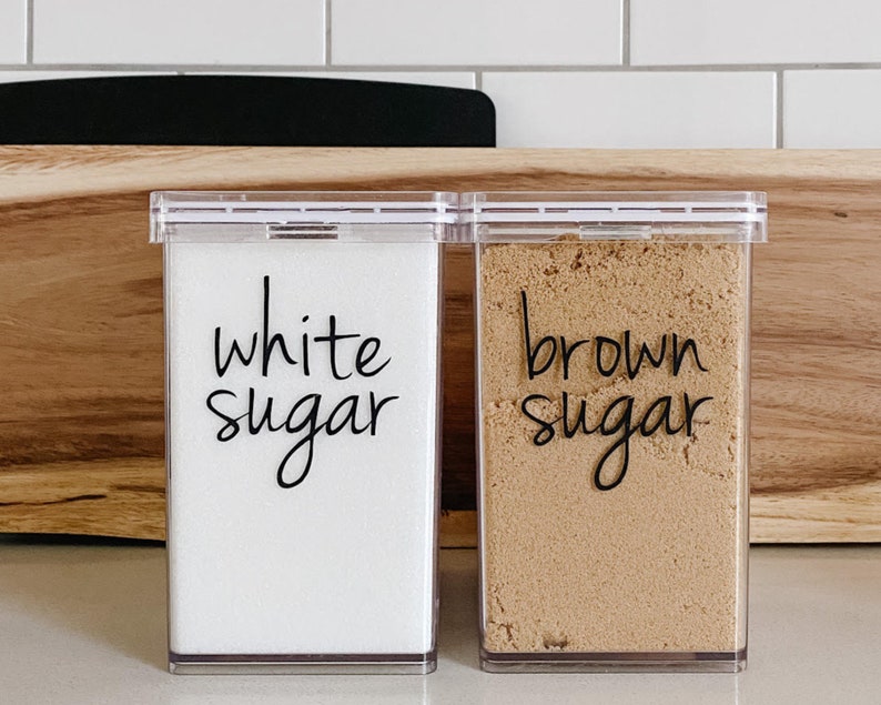 May include: Two clear plastic containers with labels that say "white sugar" and "brown sugar".