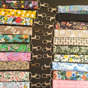 May include: A collection of colorful lanyards with various floral and patterned designs. Each lanyard is made of fabric and features a silver-colored metal clasp. The lanyards are arranged on a dark surface, showcasing a variety of colors and patterns.