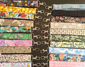 May include: A collection of colorful lanyards with various floral and patterned designs. Each lanyard is made of fabric and features a silver-colored metal clasp. The lanyards are arranged on a dark surface, showcasing a variety of colors and patterns.