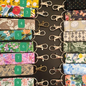 May include: A collection of fabric keychains with various patterns, including floral, geometric, and abstract designs. Each keychain features a metal clasp for easy attachment. The keychains are arranged on a dark surface.