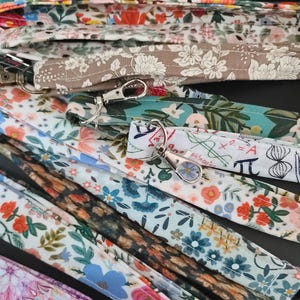 May include: A collection of patterned lanyards with metal swivel clasps. The lanyards feature various floral and botanical designs in shades of pink, blue, green, and brown. Some lanyards have a white background with mathematical symbols.