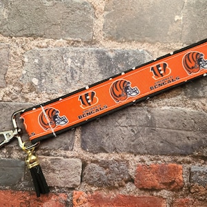 May include: A black and orange key chain with a black tassel. The key chain features a white background with black polka dots and a repeated image of a Cincinnati Bengals football helmet with the team's logo and the words "Cincinnati Bengals".