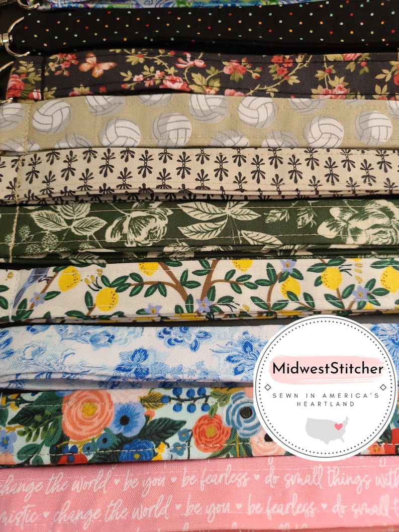 May include: A collection of fabric lanyards with various patterns, including floral, volleyballs, and polka dots. The lanyards are in a variety of colors, including black, pink, green, and blue. The text "change the world be you be fearless" is visible on a pink lanyard.