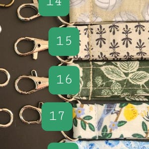 May include: A collection of fabric keychains with various patterns, including floral, polka dots, and volleyball designs. Each keychain has a metal clasp for easy attachment. The keychains are arranged on a dark surface.