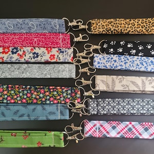 May include: A collection of lanyards with various fabric patterns, including floral, paisley, and animal print. Each lanyard has a silver-colored metal clasp. The color palette includes blue, pink, green, and neutral tones.