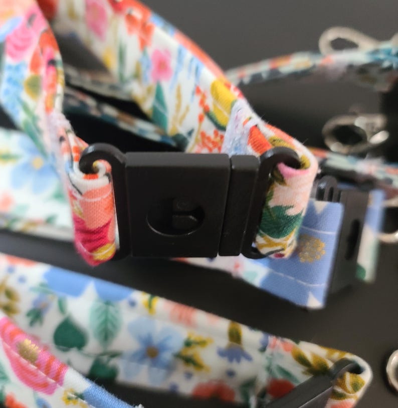 May include: Close-up of floral patterned fabric with a black plastic buckle. The fabric features a colorful design with pink, blue, and yellow flowers on a white background. The buckle is rectangular and has a curved hook.