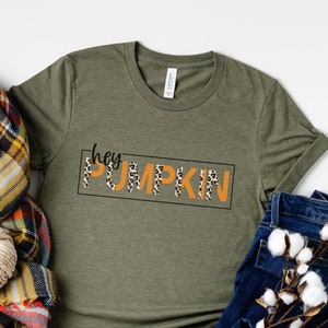 May include: A green t-shirt with the text "hey pumpkin" in black and orange letters with a leopard print pattern. The shirt is displayed on a white background with a plaid scarf and a pair of blue jeans.