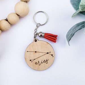 May include: A wooden keychain with a red tassel and a laser-engraved design of the Aries constellation and the word "aries".