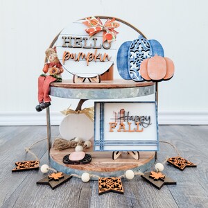 May include: A two-tiered metal stand decorated with fall-themed wooden signs. The top tier features a round sign that says "HELLO pumpkin" and a blue pumpkin with a smaller orange pumpkin. The bottom tier features a sign that says "Happy FALL" and a small white pumpkin. A wooden garland with orange and brown leaves hangs in front of the stand.