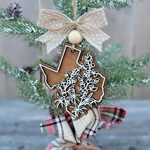 May include: A wooden ornament shaped like the state of Texas with a white outline and a design of bluebonnets inside. The ornament is hanging from a small artificial Christmas tree with a brown and white polka dot bow.