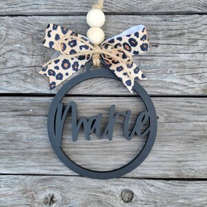 May include: Black wooden ornament with the name "Marie" cut out in a circle. The ornament is decorated with a brown and white leopard print ribbon and white wooden beads.