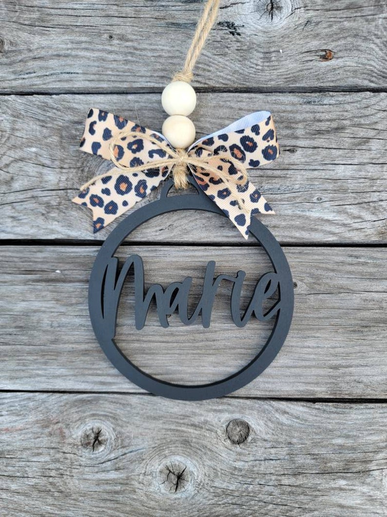 May include: Black wooden ornament with the name "Marie" cut out in a circle. The ornament is decorated with a brown and white leopard print ribbon and white wooden beads.