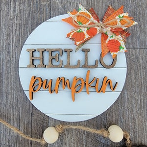 May include: A round white wooden sign with the words "hello pumpkin" in brown and orange letters. A large orange and white bow with a burlap ribbon is attached to the top of the sign.