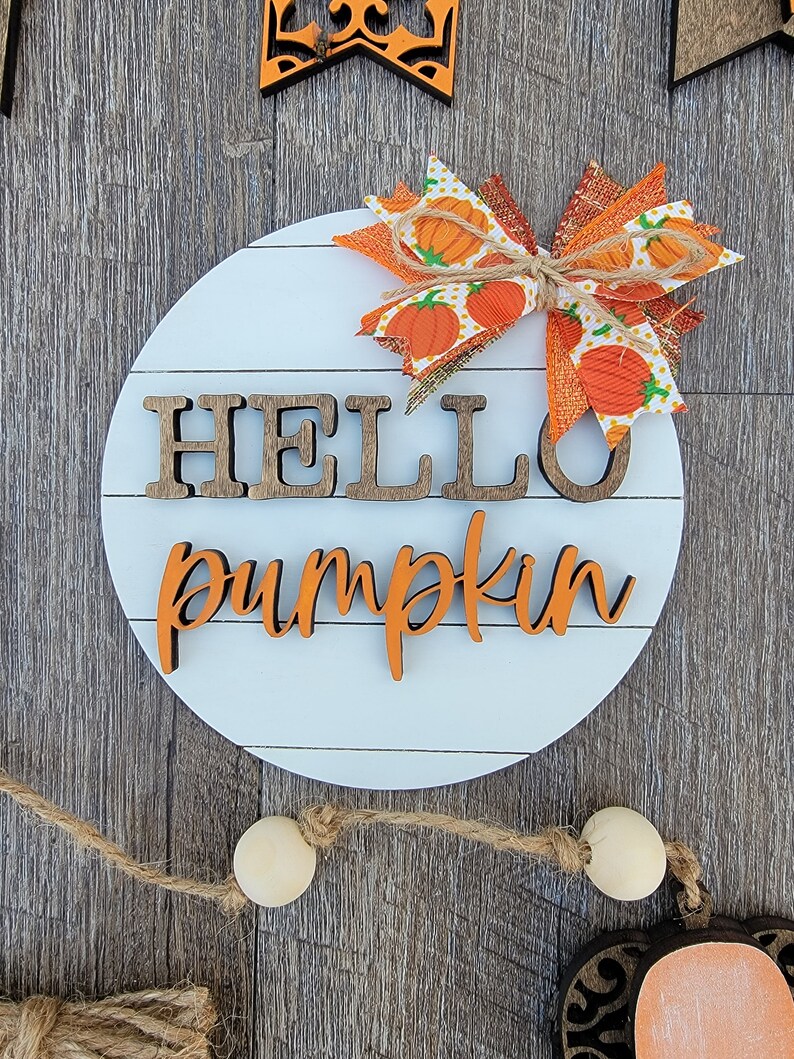 May include: A round white wooden sign with the words "hello pumpkin" in brown and orange letters. A large orange and white bow with a burlap ribbon is attached to the top of the sign.