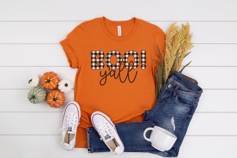 Puede incluir: Camiseta naranja con la frase "BOO! y'all" escrita en letras de cuadros negros y blancos.