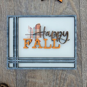May include: A wooden sign with a gray frame and a white background. The sign says "Happy Fall" in brown letters with a plaid leaf design.