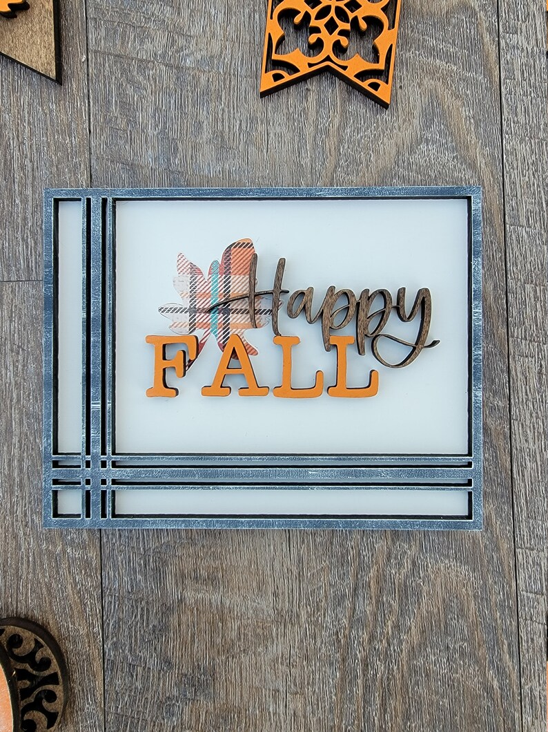 May include: A wooden sign with a gray frame and a white background. The sign says "Happy Fall" in brown letters with a plaid leaf design.