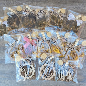 May include: A collection of 12 personalized wooden monogram ornaments, each packaged in a clear plastic bag with brown paper shred, a leopard print ribbon, and a string of wooden beads. The ornaments are in various styles, including a circle with a monogram in the center, a script monogram, and a block monogram. The ornaments are perfect for adding a personal touch to your home decor.