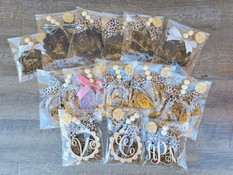May include: A collection of 12 personalized wooden monogram ornaments, each packaged in a clear plastic bag with brown paper shred, a leopard print ribbon, and a string of wooden beads. The ornaments are in various styles, including a circle with a monogram in the center, a script monogram, and a block monogram. The ornaments are perfect for adding a personal touch to your home decor.