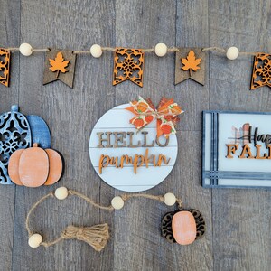 May include: A wooden sign with the text "Hello Pumpkin" in white letters on a white background. The sign is surrounded by a garland of orange and brown leaves and white beads. There is also a blue and white pumpkin with a smaller orange pumpkin on top, a sign that says "Happy Fall" in black letters on a white background, and a small orange pumpkin with a brown stem.