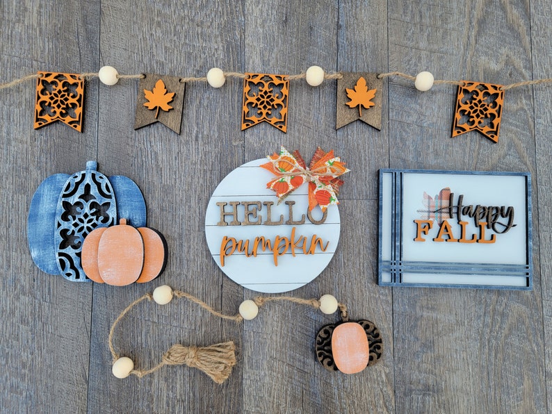 May include: A wooden sign with the text "Hello Pumpkin" in white letters on a white background. The sign is surrounded by a garland of orange and brown leaves and white beads. There is also a blue and white pumpkin with a smaller orange pumpkin on top, a sign that says "Happy Fall" in black letters on a white background, and a small orange pumpkin with a brown stem.