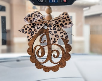 May include: A wooden car charm with a scalloped edge and a cutout letter "J" in a leopard print bow. The charm is hanging from a rearview mirror.