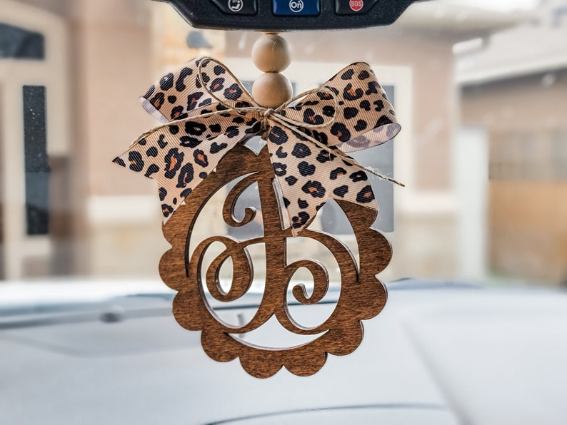May include: A wooden car charm with a scalloped edge and a cutout letter "J" in a leopard print bow. The charm is hanging from a rearview mirror.