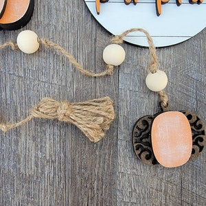 May include: A wooden pumpkin cutout with a decorative design and a light orange paint finish. The pumpkin is attached to a twine garland with wooden beads and a jute tassel.