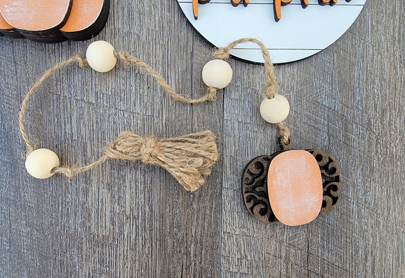 May include: A wooden pumpkin cutout with a decorative design and a light orange paint finish. The pumpkin is attached to a twine garland with wooden beads and a jute tassel.