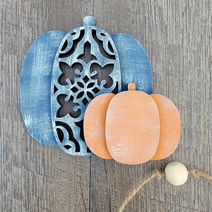 May include: Two wooden pumpkins, one blue with a cutout design and one orange, on a wood background.